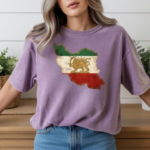 Puede incluir: Una camiseta morada con un gr&aacute;fico desgastado del mapa de Ir&aacute;n con los colores de la bandera nacional. El dise&ntilde;o incluye un emblema de le&oacute;n y sol. La camiseta es de material suave.