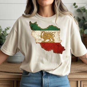 Puede incluir: Una camiseta beige con un gr&aacute;fico desgastado del mapa de Ir&aacute;n, que muestra los colores de la bandera nacional: verde, blanco y rojo. El gr&aacute;fico incluye un emblema de le&oacute;n y sol dorado. La camiseta es una prenda casual para el uso diario.
