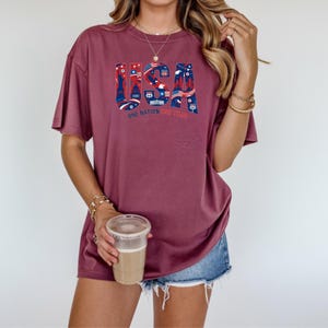 May include: A maroon t-shirt with the word "USA" in a patriotic design featuring Route 66 and other American symbols. The text "ONE NATION" and "ME TOO" are also visible on the shirt.