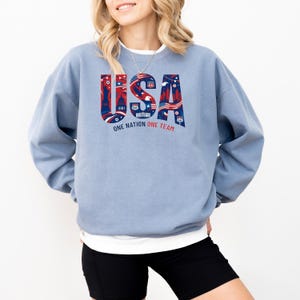 May include: A light blue sweatshirt with the word "USA" in red, white, and blue, featuring iconic American imagery. The text "ONE NATION ONE TEAM" is below the word "USA".