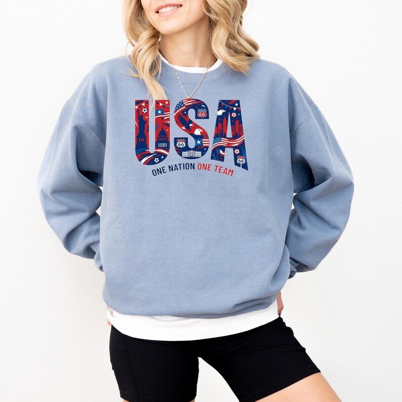 May include: A light blue sweatshirt with the word "USA" in red, white, and blue, featuring iconic American imagery. The text "ONE NATION ONE TEAM" is below the word "USA".