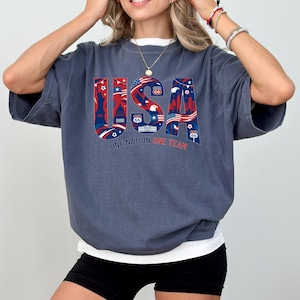 May include: A dark blue t-shirt with the word "USA" in red, white, and blue, featuring iconic American imagery. The text "ONE NATION ONE TEAM" is below the word "USA". The shirt has short sleeves and a relaxed fit.