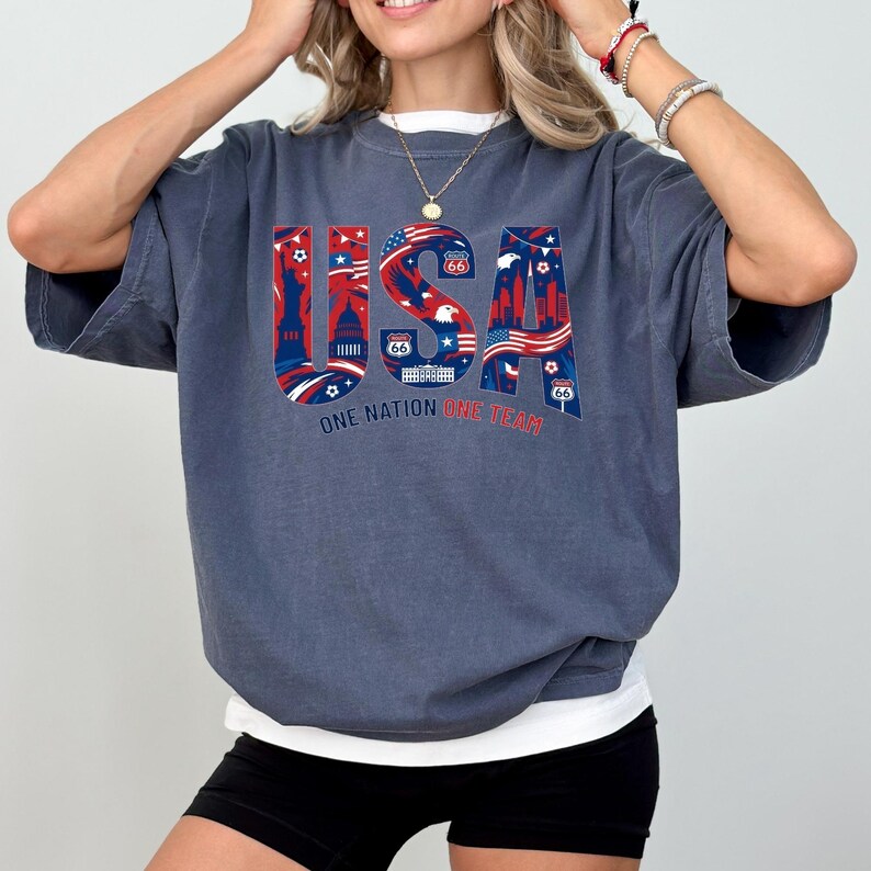 May include: A dark blue t-shirt with the word "USA" in red, white, and blue, featuring iconic American imagery. The text "ONE NATION ONE TEAM" is below the word "USA". The shirt has short sleeves and a relaxed fit.