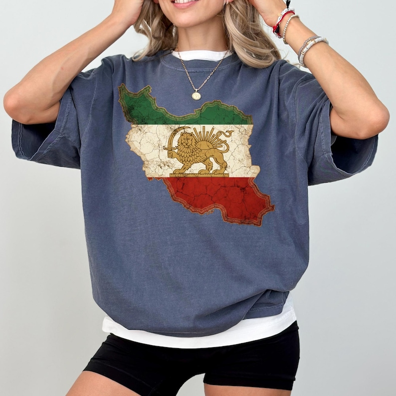 Puede incluir: Una camiseta azul con un gr&aacute;fico desgastado de la bandera iran&iacute; en un contorno de mapa. El dise&ntilde;o de la bandera incluye secciones verdes, blancas y rojas, con un emblema de le&oacute;n y sol dorado. La camiseta tiene mangas cortas y un corte holgado.