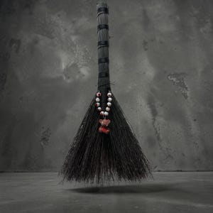 May include: A black broom with a dark handle and bristles. A beaded necklace with white beads and red accents hangs from the handle, with a small teddy bear pendant. The broom is set against a gray background.