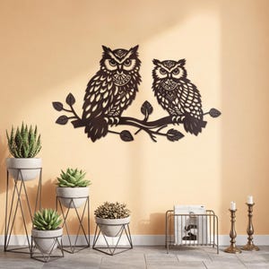 Puede incluir: Una pieza de arte mural de metal marrón oscuro con dos siluetas detalladas de búhos posados en una rama con hojas. Los búhos están colocados contra una pared de color naranja claro, con varias plantas en macetas y objetos decorativos en primer plano.