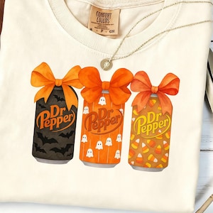 May include: Cream-colored t-shirt featuring three Dr. Pepper cans with orange bows. The cans have Halloween-themed designs: bats, ghosts, and candy corn. A gold necklace and a brown hat are also visible.