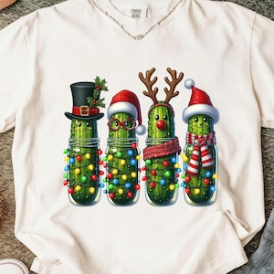 May include: White t-shirt featuring a festive design of four anthropomorphic pickles in glass jars, each adorned with Christmas accessories like hats, antlers, and scarves. The pickles are decorated with colorful string lights.