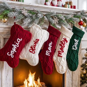 May include: Five knit Christmas stockings hanging from a mantelpiece. Two are red, two are white, and one is dark green. The stockings feature cable knit patterns and embroidered names: Sophia, Olivia, mom, molly, and Dad. The two red stockings are shaped like dog bones.