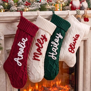 May include: Four cable-knit Christmas stockings hanging in a row. The first is red with white text reading Amelia, the second is white with red text reading molly, the third is green with white text reading Hailey, and the fourth is white with red text reading Olivia.