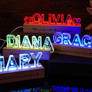 Peut inclure: Enseignes lumineuses avec des noms et des motifs floraux. Les noms "OLIVIA", "DIANA", "GRACE" et "MARY" sont affichés en lettres claires, chacune avec une couleur et un motif floral uniques. Les enseignes reposent sur des socles en bois avec des citations inspirantes.