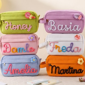 Puede incluir: Seis estuches de lápices de pana de varios colores, cada uno con un nombre bordado en la parte delantera. Los colores incluyen verde, morado, rosa, blanco, azul y naranja. Cada estuche tiene cierre de cremallera y un adorno floral o en espiral.