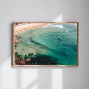 Könnte beinhalten: Luftaufnahme einer Strandszene in einem Holzrahmen. Das Bild zeigt einen Sandstrand, türkisfarbenes Wasser und dunkelgrüne, mit Algen bedeckte Felsen. Das Wasser geht von klar zu tiefblau über, mit kleinen Wellen.