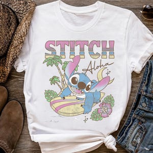 K&ouml;nnte beinhalten: Wei&szlig;es T-Shirt mit einer farbenfrohen Grafik von Stitch aus Lilo & Stitch. Das Design enth&auml;lt das Wort "STITCH" in einem Retro-Schriftzug, das Wort "Aloha" und eine Cartoon-Figur von Stitch beim Surfen auf einem Surfbrett. Das T-Shirt ist von tropischen Elementen umgeben.