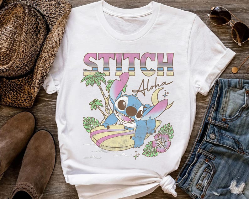 K&ouml;nnte beinhalten: Wei&szlig;es T-Shirt mit einer farbenfrohen Grafik von Stitch aus Lilo & Stitch. Das Design enth&auml;lt das Wort "STITCH" in einem Retro-Schriftzug, das Wort "Aloha" und eine Cartoon-Figur von Stitch beim Surfen auf einem Surfbrett. Das T-Shirt ist von tropischen Elementen umgeben.
