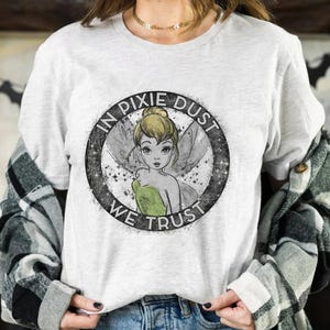 Op de afbeelding: Lichtgrijs t-shirt met een cirkelvormige afbeelding met de tekst "IN PIXIE DUST WE TRUST". De afbeelding toont een fee met blond haar en groene kleding. Dit is een casual t-shirt.