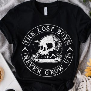 May include: Black t-shirt with a white skull graphic and the text "THE LOST BOYS NEVER GROW UP" in a circular design. The shirt is made of a soft material and has short sleeves.