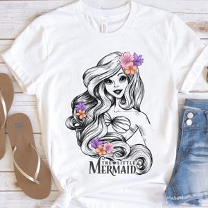 May include: White t-shirt featuring a black and white illustration of a mermaid with long flowing hair adorned with flowers. The text "THE LITTLE MERMAID" is below the illustration. Brown sandals and denim shorts are also visible.