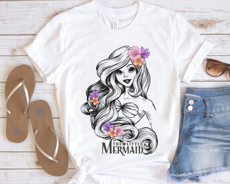May include: White t-shirt featuring a black and white illustration of a mermaid with long flowing hair adorned with flowers. The text "THE LITTLE MERMAID" is below the illustration. Brown sandals and denim shorts are also visible.