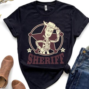 Puede incluir: Camiseta negra con un dise&ntilde;o de sheriff de dibujos animados dentro de una insignia en forma de estrella. El sheriff lleva sombrero de vaquero y chaleco. La palabra "SHERIFF" est&aacute; impresa en granate debajo.