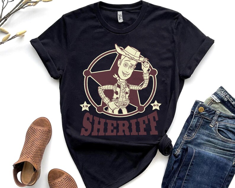 Puede incluir: Camiseta negra con un dise&ntilde;o de sheriff de dibujos animados dentro de una insignia en forma de estrella. El sheriff lleva sombrero de vaquero y chaleco. La palabra "SHERIFF" est&aacute; impresa en granate debajo.
