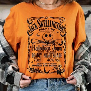 May include: Orange long-sleeve shirt with a black graphic featuring Jack Skellington's name and a Halloween-themed design. The text includes "Halloween Town" and "Deadly Nightshade". The shirt also has "70cl" and "40% Vol." printed on it.