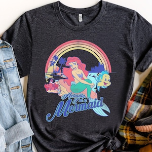 Pode incluir: Camiseta cinza escuro com um gr&aacute;fico colorido de uma sereia com cabelo ruivo, uma cauda verde e um peixe amarelo. O design inclui um arco-&iacute;ris e o texto "Actually I'm a Mermaid."