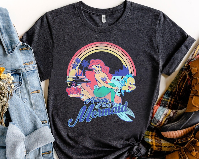 Pode incluir: Camiseta cinza escuro com um gr&aacute;fico colorido de uma sereia com cabelo ruivo, uma cauda verde e um peixe amarelo. O design inclui um arco-&iacute;ris e o texto "Actually I'm a Mermaid."