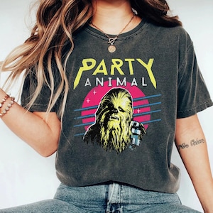 May include: Dark grey t-shirt with a graphic featuring the text "PARTY ANIMAL" in yellow above an image of Chewbacca. The image includes a pink circle, blue lines, and a retro design.