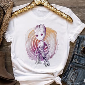 May include: White t-shirt featuring a graphic of a Groot character from Guardians of the Galaxy. The design shows Groot standing in front of a circular, watercolor-style background with orange, purple, and pink hues. The shirt is displayed on a gold sequined hanger.