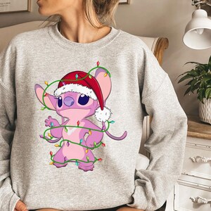 May include: A light gray sweatshirt featuring a cartoon character, Stitch, wearing a Santa hat and wrapped in Christmas lights. The character is pink with large blue eyes and is smiling. The lights are green with colorful bulbs.