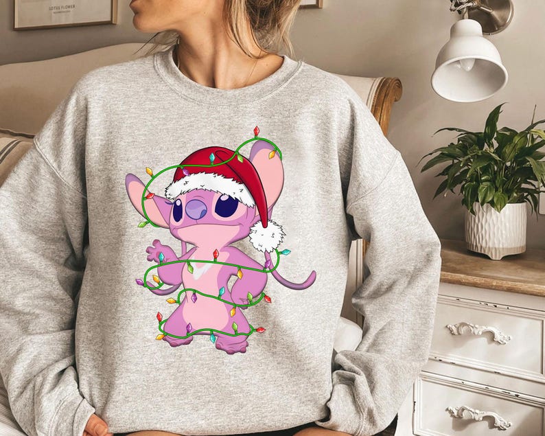 May include: A light gray sweatshirt featuring a cartoon character, Stitch, wearing a Santa hat and wrapped in Christmas lights. The character is pink with large blue eyes and is smiling. The lights are green with colorful bulbs.