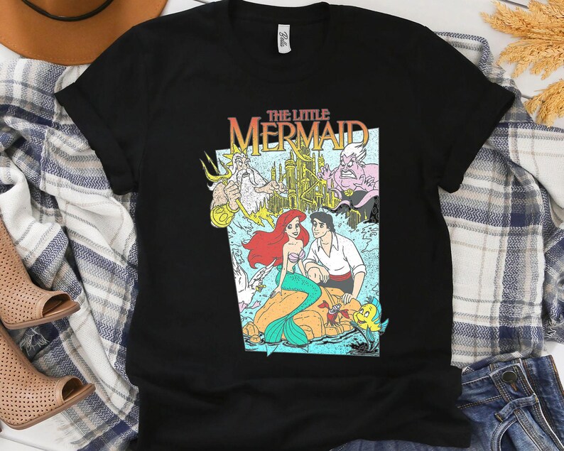 Puede incluir: Camiseta negra con un gr&aacute;fico colorido de La Sirenita de Disney. El dise&ntilde;o incluye a Ariel, el Pr&iacute;ncipe Eric y otros personajes, con el t&iacute;tulo en una fuente retro. Una camiseta cl&aacute;sica para los fans de la pel&iacute;cula animada.