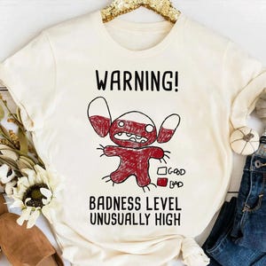 Puede incluir: Camiseta color crema con una ilustraci&oacute;n de personaje de dibujos animados rojo y blanco y el texto "WARNING! BADNESS LEVEL UNUSUALLY HIGH." El dise&ntilde;o incluye una escala "bueno" y "malo". La camiseta se muestra con vaqueros y objetos decorativos.