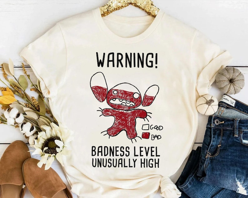 Puede incluir: Camiseta color crema con una ilustraci&oacute;n de personaje de dibujos animados rojo y blanco y el texto "WARNING! BADNESS LEVEL UNUSUALLY HIGH." El dise&ntilde;o incluye una escala "bueno" y "malo". La camiseta se muestra con vaqueros y objetos decorativos.