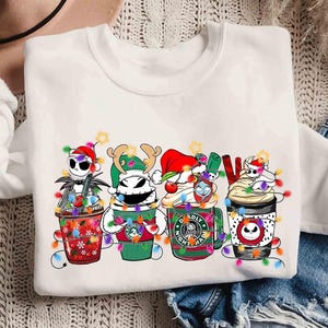 May include: White crew neck sweatshirt featuring a colorful graphic of Nightmare Before Christmas characters as coffee cups, each decorated with Christmas lights and festive hats. The word "NOEL" is also in the design.