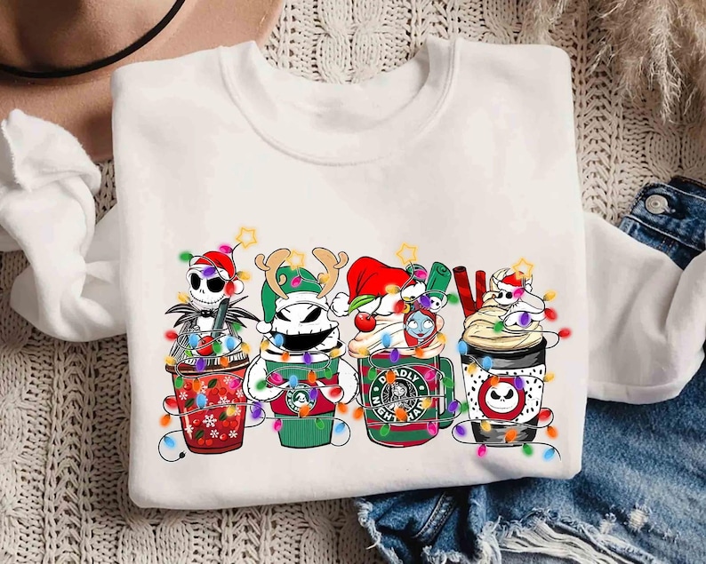 May include: White crew neck sweatshirt featuring a colorful graphic of Nightmare Before Christmas characters as coffee cups, each decorated with Christmas lights and festive hats. The word "NOEL" is also in the design.