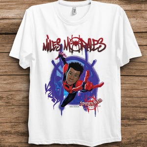 May include: A white short-sleeved t-shirt featuring a graphic of Miles Morales in a red and black suit. The text "MILES MORALES" appears in a red graffiti style above, with "SPIDER-MAN INTO THE SPIDER-VERSE" printed in the lower right corner.