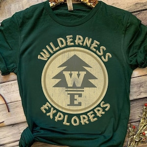 Może przedstawiać: Ciemnozielona koszulka z okrągłą grafiką przedstawiającą stylizowaną sosnę i napis "WILDERNESS EXPLORERS". Projekt jest w wyblakłym beżowym kolorze, a koszulka ma podwinięte rękawy.