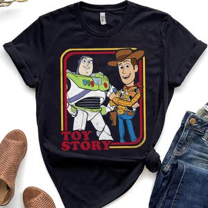 Puede incluir: Camiseta negra con un gr&aacute;fico retro de Buzz Lightyear y Woody de Toy Story. El dise&ntilde;o est&aacute; enmarcado en un borde rojo y amarillo, con las palabras "TOY STORY" en letras rojas en la parte inferior.