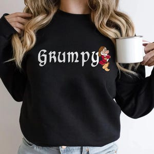 May include: Black sweatshirt with the word "Grumpy" in white script, accompanied by a cartoon image of Grumpy from Snow White. The sweatshirt is a crew neck style.