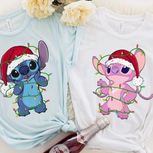 May include: Two t-shirts, one light blue and one white, each featuring a cartoon character wearing a Santa hat and wrapped in Christmas lights. The blue character is blue, and the white shirt features a pink character.