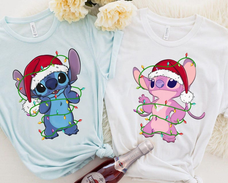 May include: Two t-shirts, one light blue and one white, each featuring a cartoon character wearing a Santa hat and wrapped in Christmas lights. The blue character is blue, and the white shirt features a pink character.