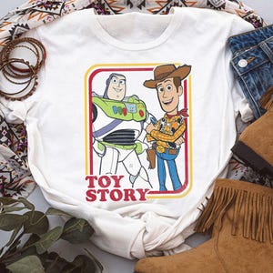 Puede incluir: Camiseta blanca con un gr&aacute;fico de Buzz Lightyear y Woody de Toy Story, enmarcado en rojo y amarillo. Las palabras "TOY STORY" est&aacute;n impresas en rojo. La camiseta se combina con vaqueros y botas marrones con flecos.