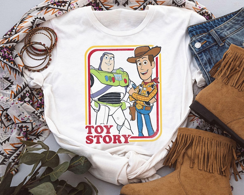 Puede incluir: Camiseta blanca con un gr&aacute;fico de Buzz Lightyear y Woody de Toy Story, enmarcado en rojo y amarillo. Las palabras "TOY STORY" est&aacute;n impresas en rojo. La camiseta se combina con vaqueros y botas marrones con flecos.