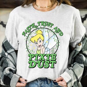 Op de afbeelding: Lichtgrijs t-shirt met een cartoonafbeelding van Tinkerbell in een groene cirkel, en de woorden "Faith Trust And Pixie Dust" in groene letters. De fee draagt een groene jurk en heeft blond haar.