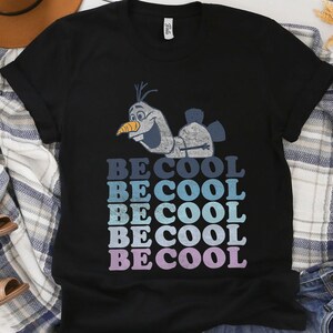 Puede incluir: Camiseta negra con una imagen de dibujos animados de Olaf de Frozen. La camiseta tiene las palabras "BE COOL" en un dise&ntilde;o apilado, con cada palabra en un tono diferente de azul y morado. La camiseta est&aacute; hecha de un material suave.