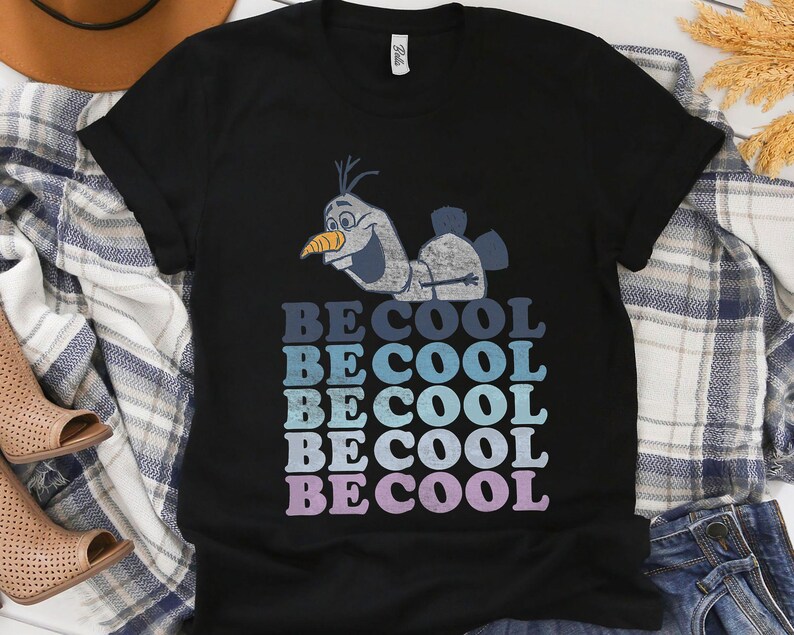 Puede incluir: Camiseta negra con una imagen de dibujos animados de Olaf de Frozen. La camiseta tiene las palabras "BE COOL" en un dise&ntilde;o apilado, con cada palabra en un tono diferente de azul y morado. La camiseta est&aacute; hecha de un material suave.