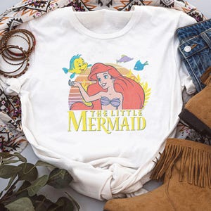 May include: White t-shirt featuring a colorful graphic of Ariel from The Little Mermaid, with the text "THE LITTLE MERMAID". The design includes Ariel, Flounder, and other sea creatures against a sunset backdrop. The shirt is paired with denim jeans and brown boots.