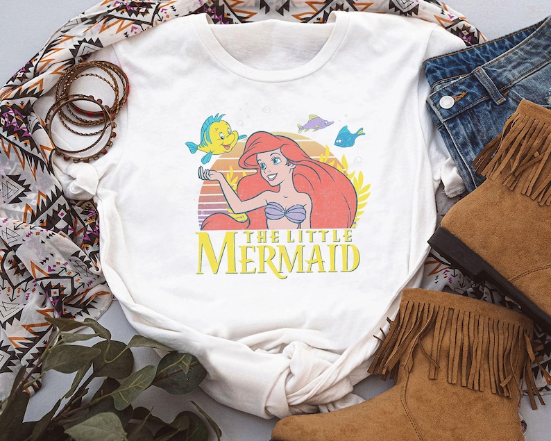 May include: White t-shirt featuring a colorful graphic of Ariel from The Little Mermaid, with the text "THE LITTLE MERMAID". The design includes Ariel, Flounder, and other sea creatures against a sunset backdrop. The shirt is paired with denim jeans and brown boots.
